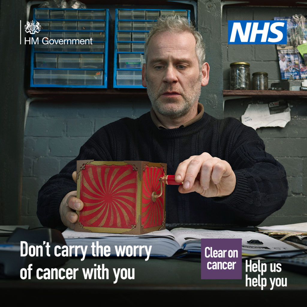 A middle-aged person sitting with a red jack-in-the-box toy on the table across from them. The person is turning the handle of the red-jack-in-the-box and has a concerned expression on their face. The text reads: ‘Don’t carry the worry of cancer with you’. The NHS logo is in the top right hand corner.
